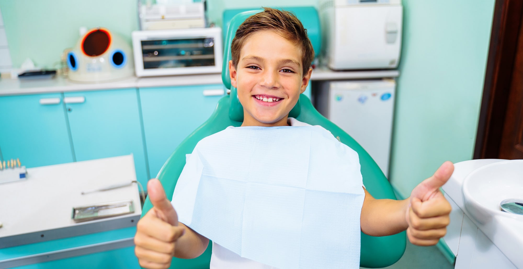 A happy child sits in an orthodontist chair while giving two thumbs up